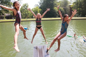 Three summer camp girls jump off the diving board at summer camp.