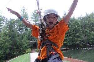 A summer camp girl in an orange shirt rides the zip line with hands stretched out.