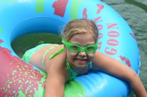 A girl in a green bathing suit and green goggles smiles from inside an innertube.