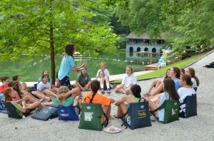 Girls sit in a circle with McLeod Lodge and the swim lake in the background.