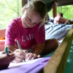 A camper in a pink Illahee t-shirt sits Indian-style on her top bunk writing a letter home.