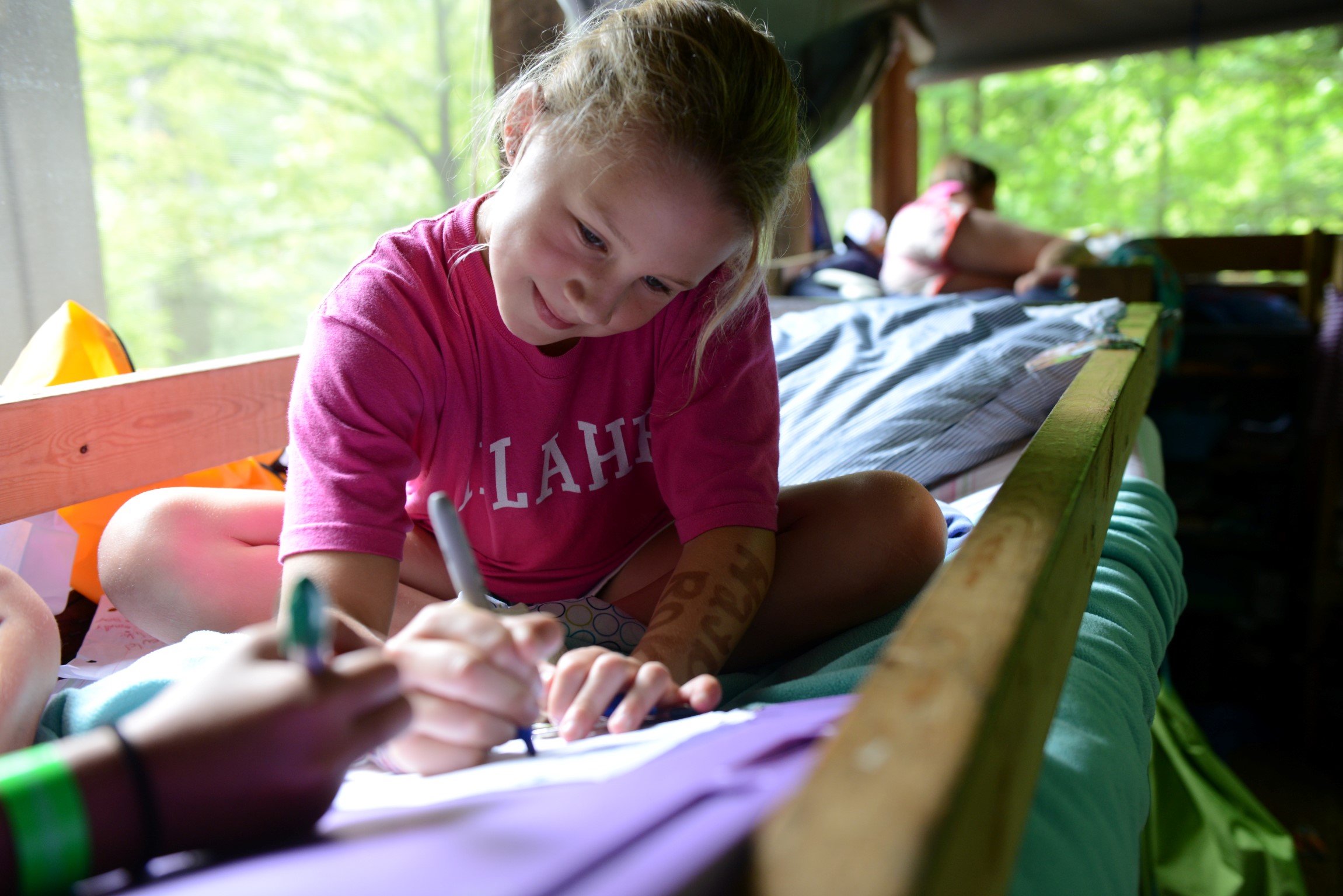 A camper in a pink Illahee t-shirt sits Indian-style on her top bunk writing a letter home.