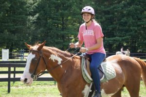 An Illahee camper in a pink Illahee t-shirt smiles form her perch above a horse.